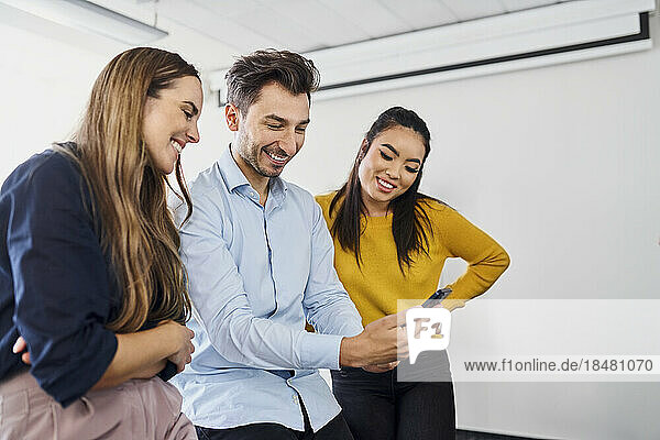 Happy businessman sharing smart phone with colleagues standing at office