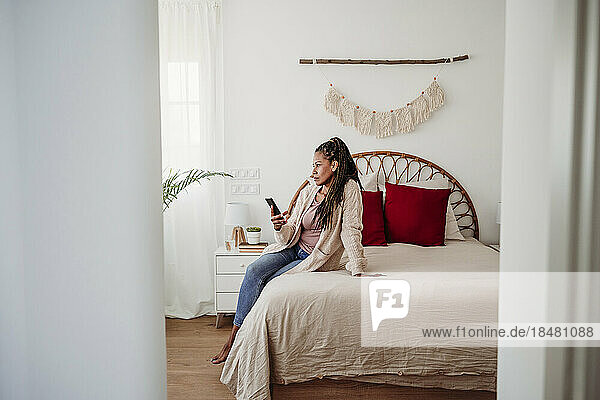 Nachdenkliche Frau sitzt mit Smartphone zu Hause im Bett
