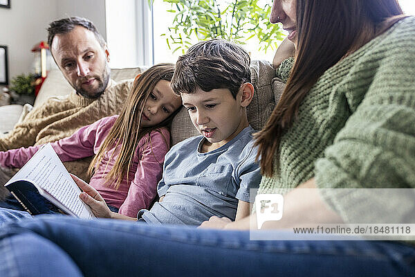 Boy reading book with family sitting on sofa at home