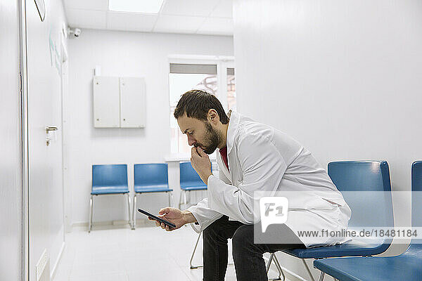 Worried doctor using smartphone sitting on chair in corridor