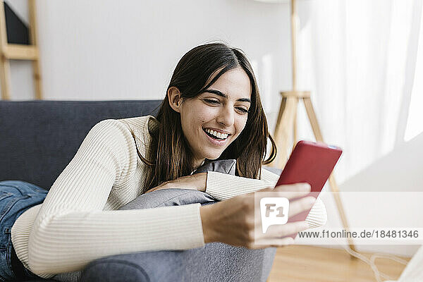 Happy young woman using smart phone lying on sofa at home