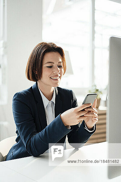 Glückliche Geschäftsfrau sitzt am Schreibtisch im Büro und nutzt ihr Smartphone