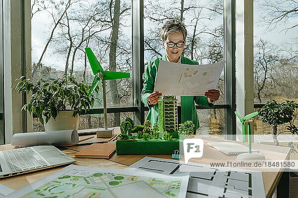 Businesswoman holding blueprint with biophilic architecture and wind turbine models at table in office