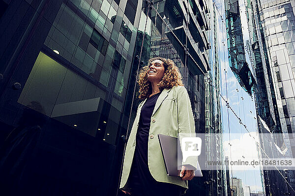 Glückliche Geschäftsfrau mit Laptop in der Nähe eines modernen Glasgebäudes