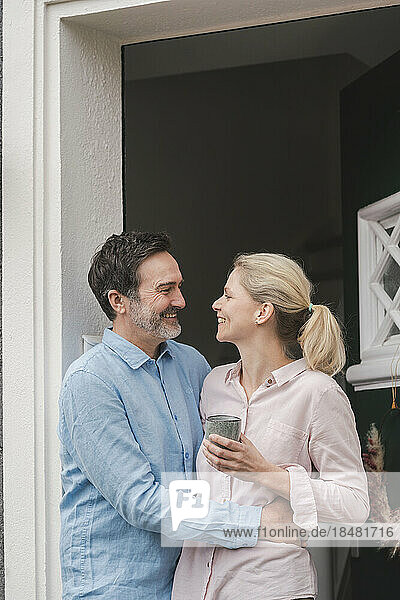 Happy man embracing wife with cup at doorway of house