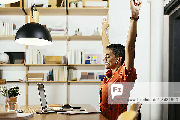 Happy freelancer sitting with arms raised in front of laptop at table