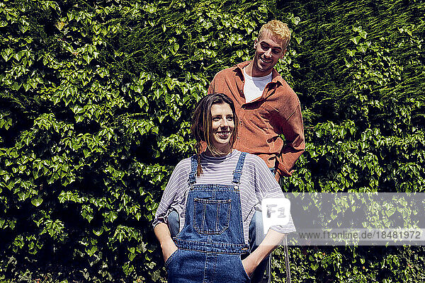 Smiling woman with hands in pockets standing by boyfriend near hedge