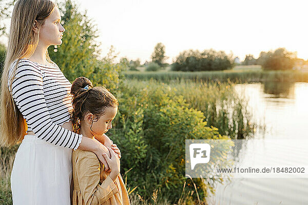 Thoughtful girl standing with sister by lake at sunset