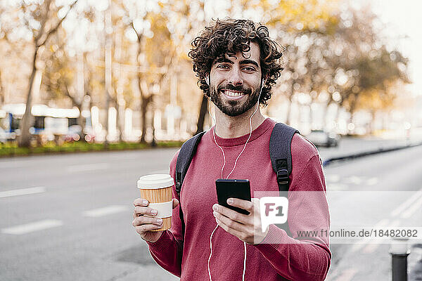 Happy young man with mobile phone and coffee cup standing at footpath