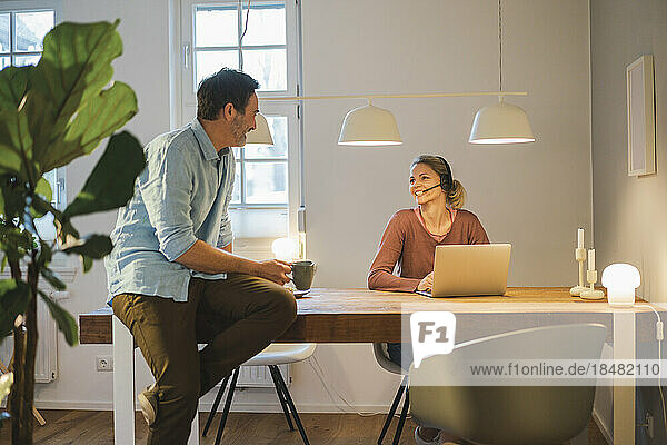 Glückliche Frau mit Headset im Gespräch mit einem Mann  der im Heimbüro am Schreibtisch sitzt