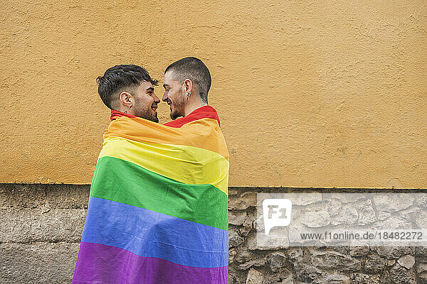 Happy gay couple covered in rainbow flag by wall