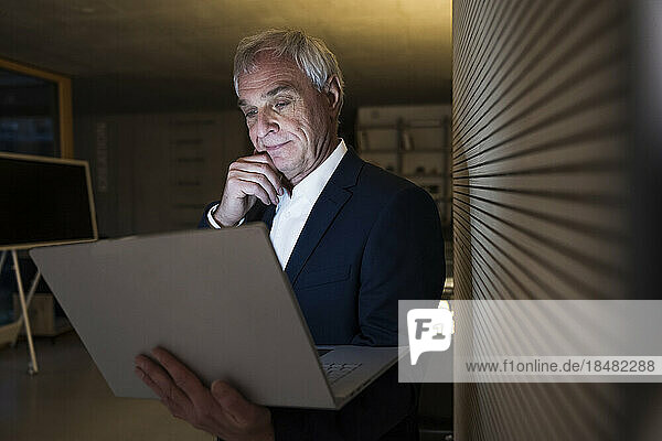 Nachdenklicher Geschäftsmann mit der Hand am Kinn  der im Büro auf den Laptop schaut