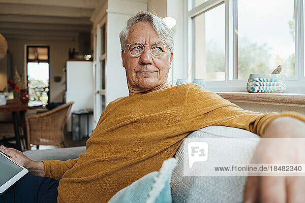 Nachdenklicher älterer Mann sitzt mit Tablet-PC auf dem Sofa im heimischen Wohnzimmer