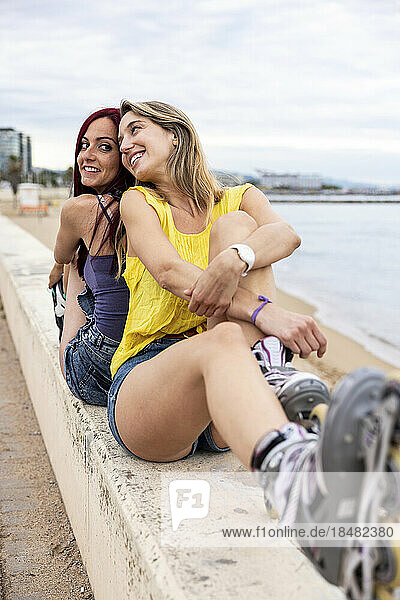 Smiling friends wearing inline skates sitting back to back at promenade