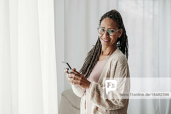 Smiling freelancer with smart phone standing in front of curtains