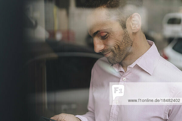 Smiling mature businessman using smart phone seen through glass