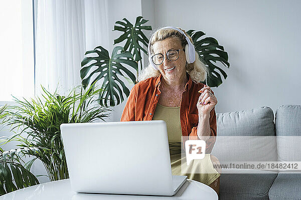 Smiling woman enjoying music through headphones and laptop sitting at home