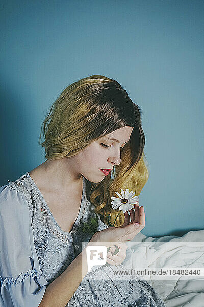 Schöne blonde Frau  die eine Blume in der Nähe einer blauen Wand betrachtet
