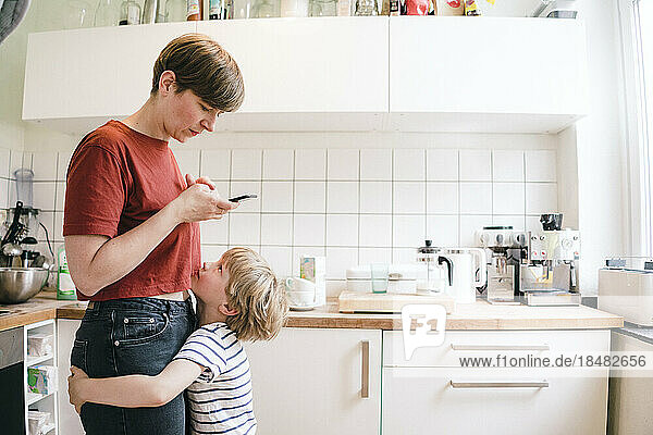 Boy embracing mother using smart phone standing in kitchen