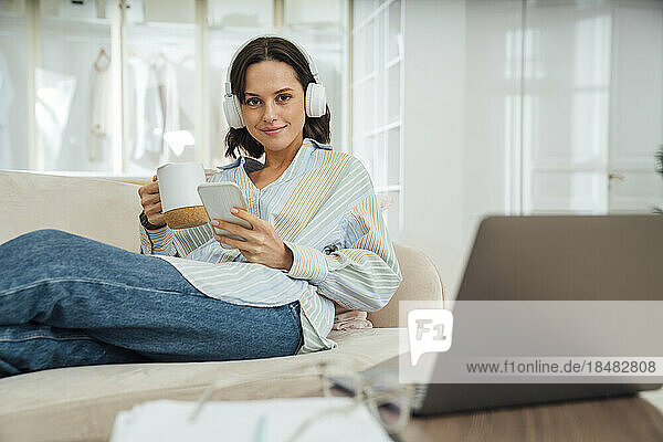 Smiling freelancer wearing wireless headphones sitting with smart phone and coffee cup on sofa at home