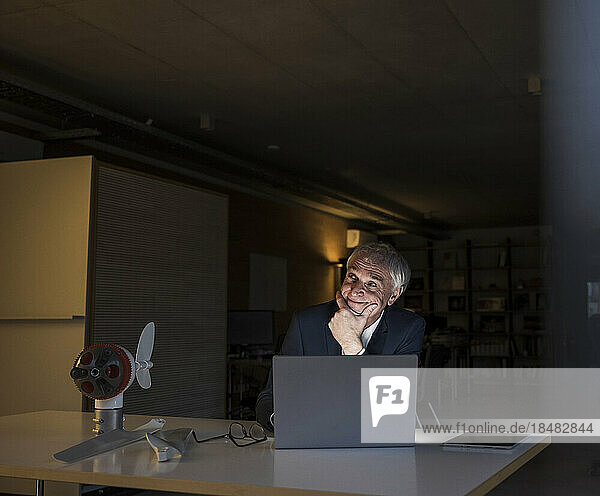 Lächelnder Geschäftsmann mit der Hand am Kinn sitzt am Schreibtisch im Büro