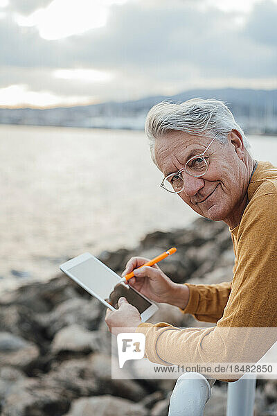 Smiling senior man with tablet PC and digitized pen leaning on railing