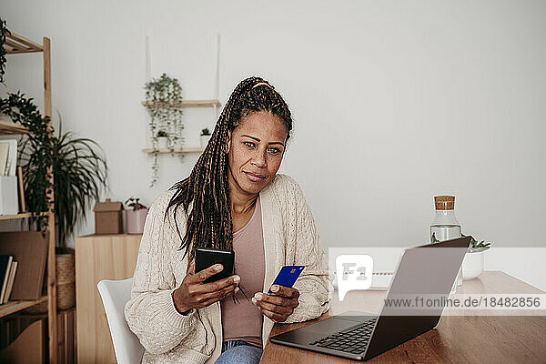 Mature freelancer with credit card and smart phone sitting at desk