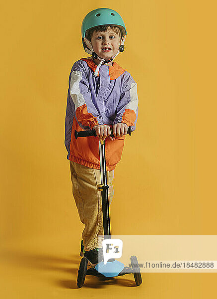 Smiling boy wearing helmet standing with push scooter against yellow background