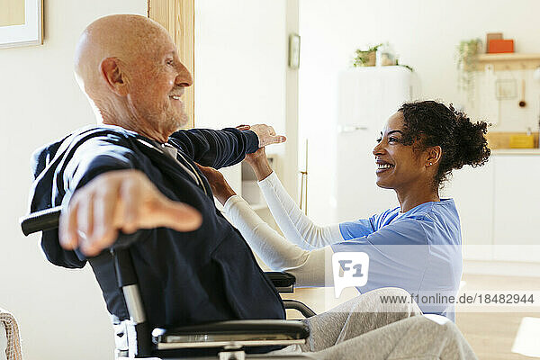 Smiling physical therapist examining senior man stretching arms at home