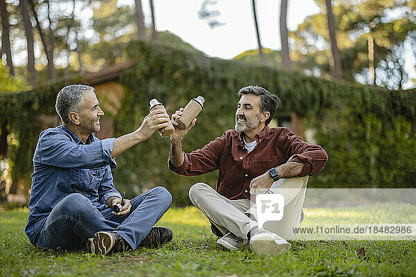 Mature friends sitting in grass clinking reusable bottles