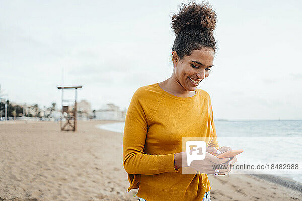 Smiling young woman text messaging using smart phone at shore on beach