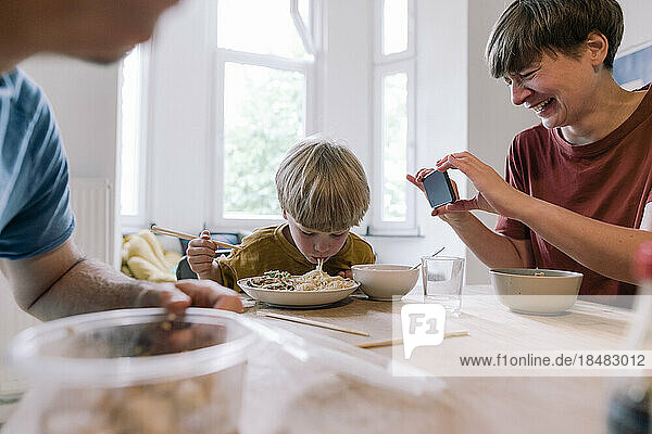 Happy woman photographing son eating food at home