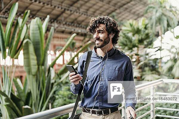 Lächelnder Geschäftsmann mit In-Ear-Kopfhörern steht mit Smartphone neben Pflanzen