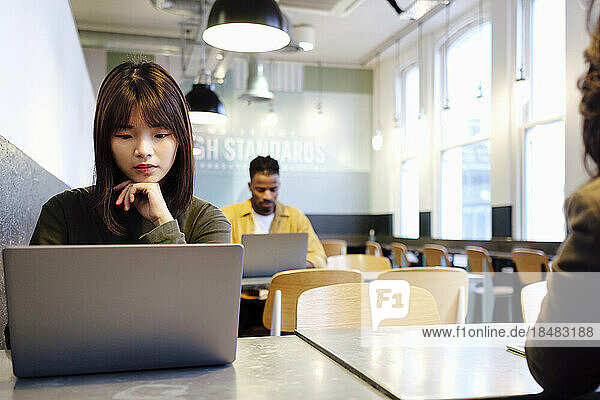 Junge Geschäftsfrau mit der Hand am Kinn und Laptop in der Cafeteria