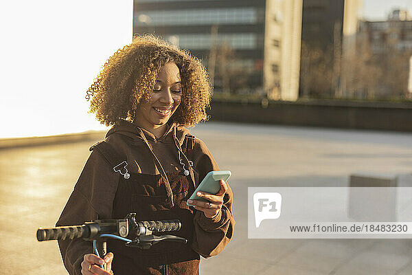 Smiling woman with push scooter using mobile phone at sunset