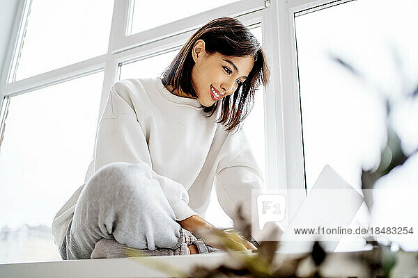 Happy woman using laptop on window sill at home