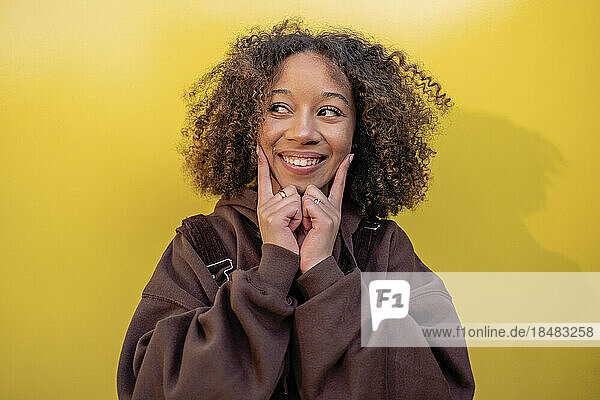 Smiling woman with curly hair touching face in front of yellow wall