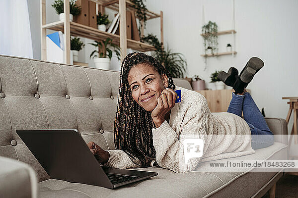 Smiling freelancer lying on sofa with credit card and laptop