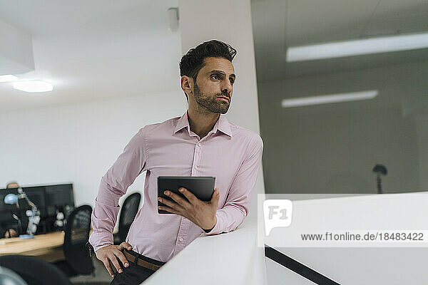 Thoughtful businessman with tablet PC leaning on wall in office