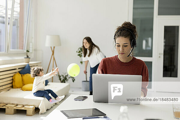 Young woman working from home while nanny is playing with daughter