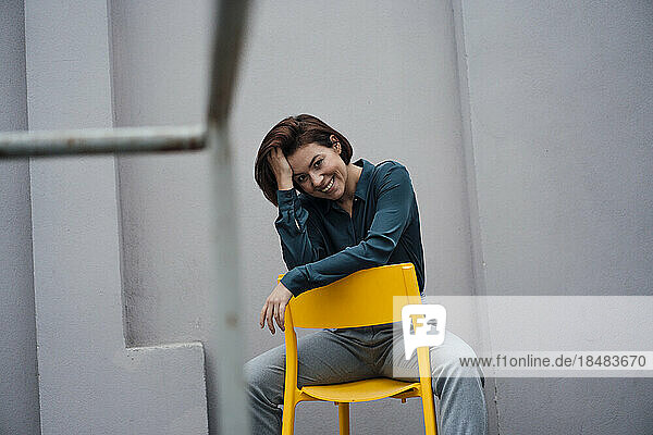 Lächelnde junge Geschäftsfrau sitzt auf einem Stuhl vor einer grauen Wand