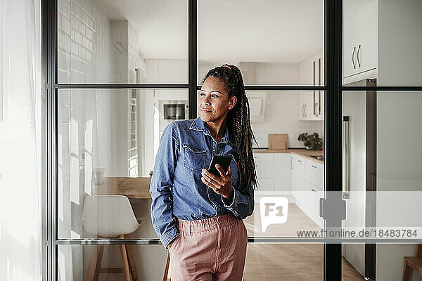 Thoughtful woman standing with mobile phone at home