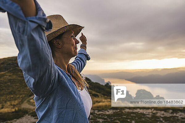 Carefree mature woman with arms raised standing under sky at sunset
