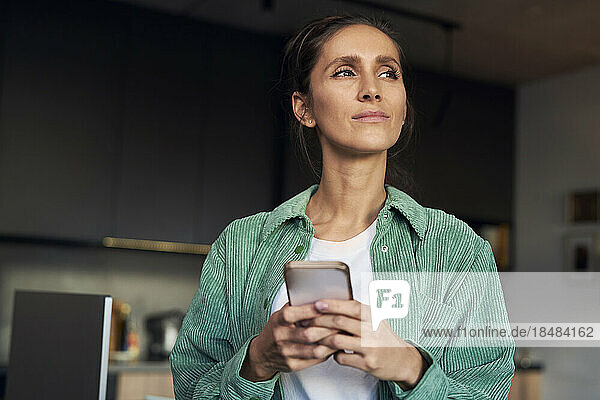 Thoughtful woman with smart phone standing at home