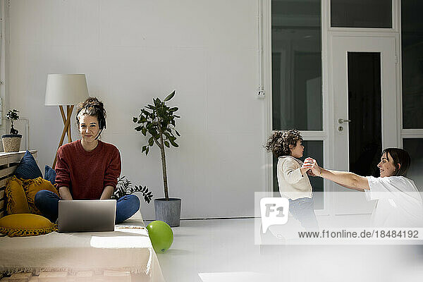 Young woman working from home while nanny is playing with daughter