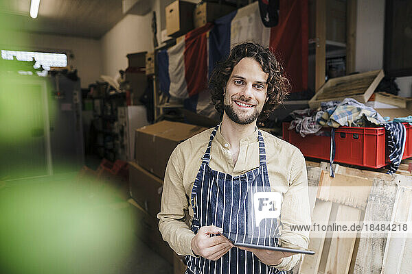 Happy owner wearing apron standing with tablet PC in storage room