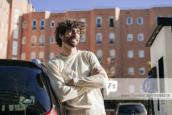 Lächelnder junger Mann stützt sich an der Fahrzeugladestation auf ein Elektroauto