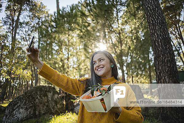 Lächelnde reife Frau hält eine Kiste mit Mandarinen in der Hand und macht ein Selfie