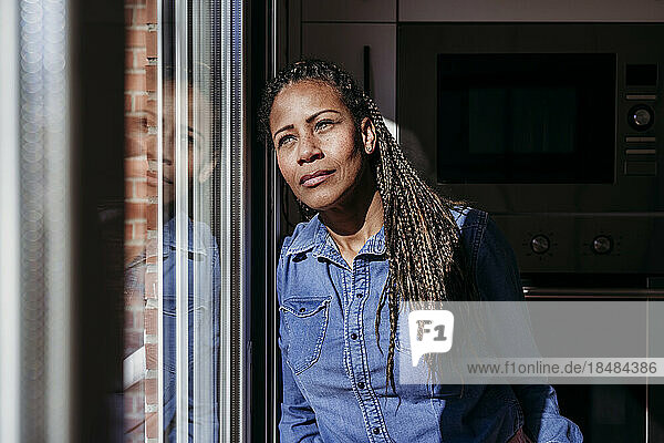 Thoughtful mature woman leaning on glass door at home