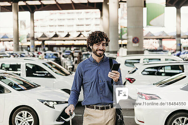 Smiling young businessman with mobile phone standing in front of cars